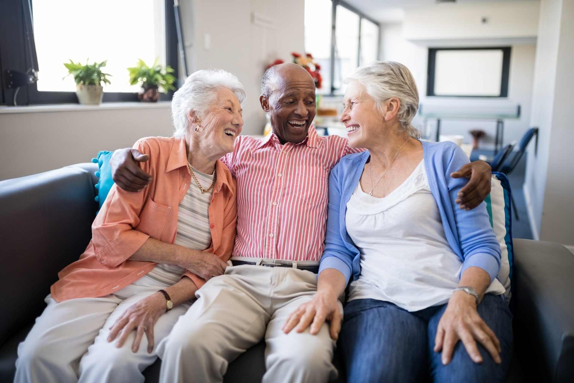 Three smiling senior adults sitting together on a couch in a bright senior living community, sharing a joyful conversation and enjoying companionship.